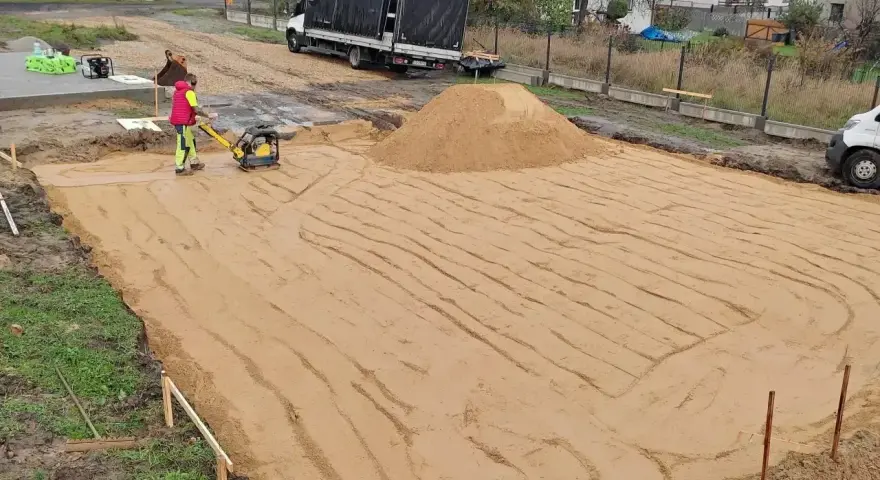 Pracownik zagęszcza piasek zagęszczarką. To kluczowy etap wymiany gruntu pod fundament, zapewniający stabilność przyszłego budynku.