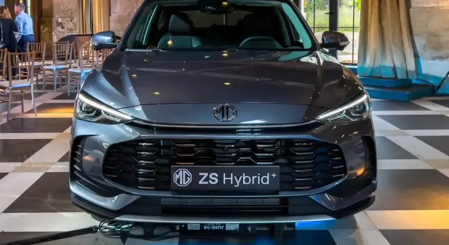 Szary MG ZS Hybrid+ prezentuje się elegancko na tle wnętrza z zasłonami i krzesłami.