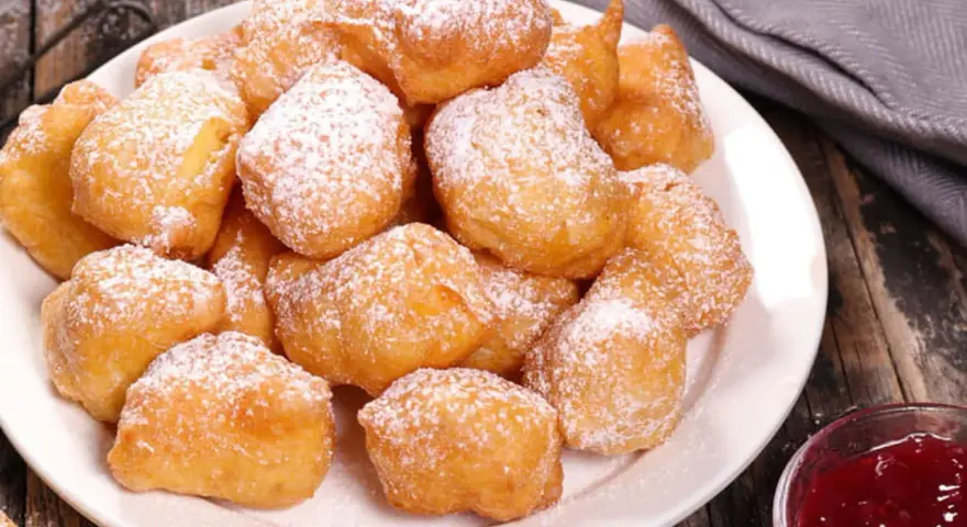 Une assiette de beignets banane saupoudrés de sucre glace, prêts à être dégustés avec une confiture de fruits rouges.