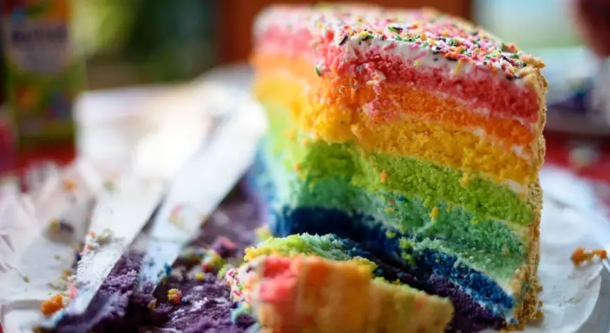 Ein Stück bunter **Regenbogen Kuchen** mit bunten Streuseln, das auf einem Teller liegt. Daneben liegen Krümel und ein Messer.