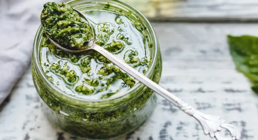 Un vasetto di pesto genovese fatto in casa con frullatore, pronto per essere gustato. Un cucchiaino d'argento raccoglie un po' di questa delizia verde.