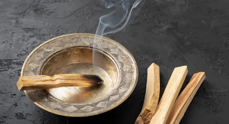 Un palo santo arde en un cuenco de metal, liberando humo blanco. A su lado, tres palos de palo santo sin quemar. Se puede quemar palo santo de noche para purificar el ambiente.