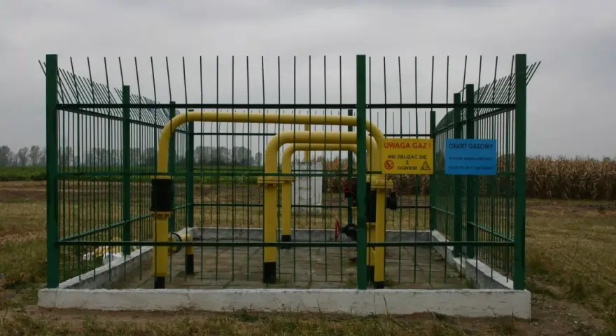 Żółty gazociąg wysokiego ciśnienia na działce, otoczony zielonym ogrodzeniem. Tablice ostrzegają przed gazem i ogniem.