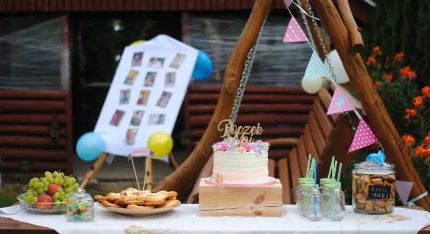 Na stole na roczek kładzie się tort, owoce, ciasteczka i słodkości. Dekoracje tworzą girlandy i zdjęcia.