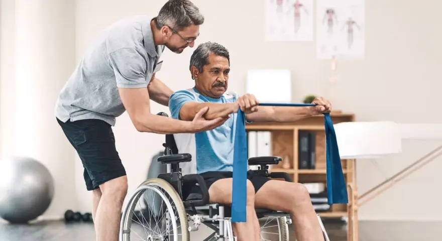 Ein Mann im Rollstuhl trainiert mit einem Therapeuten, der ihm hilft, die Widerstandsbandübungen auszuführen. Die Physiotherapeuten unterstützen ihn bei seiner Genesung.