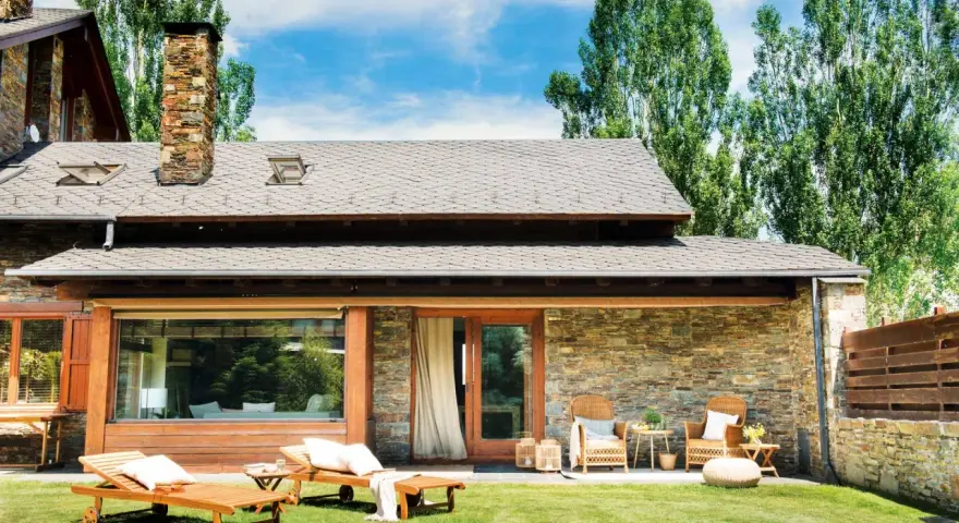 Fachadas de casas de pueblo modernas con piedra y madera, jardín con tumbonas y zona de estar exterior.