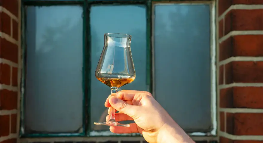 Ein Glas bernsteinfarbener Whisky wird vor einem Fenster gehalten. Der Alkoholgehalt ist bei dieser Verkostung entscheidend.