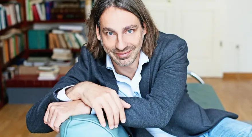 Richard David Precht sitzt lächelnd auf einem türkisfarbenen Sofa, im Hintergrund ein Bücherregal.