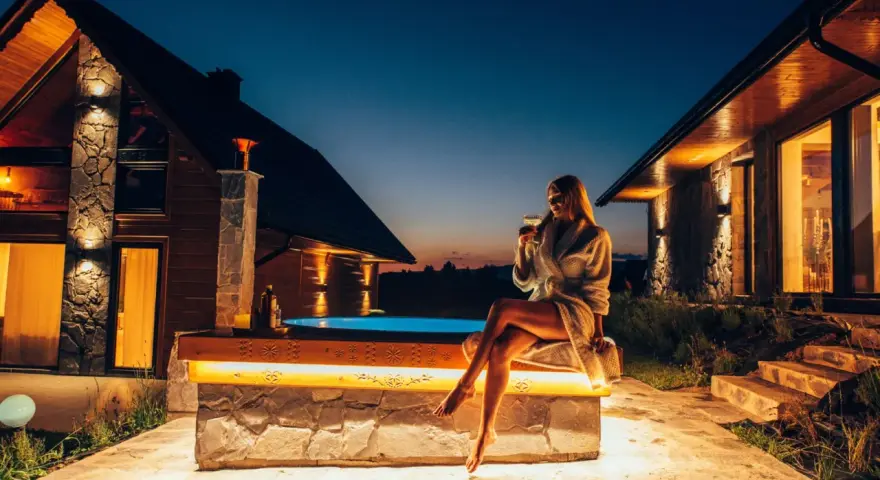 Kobieta w szlafroku z drinkiem siedzi przy jacuzzi przed górskim domkiem.