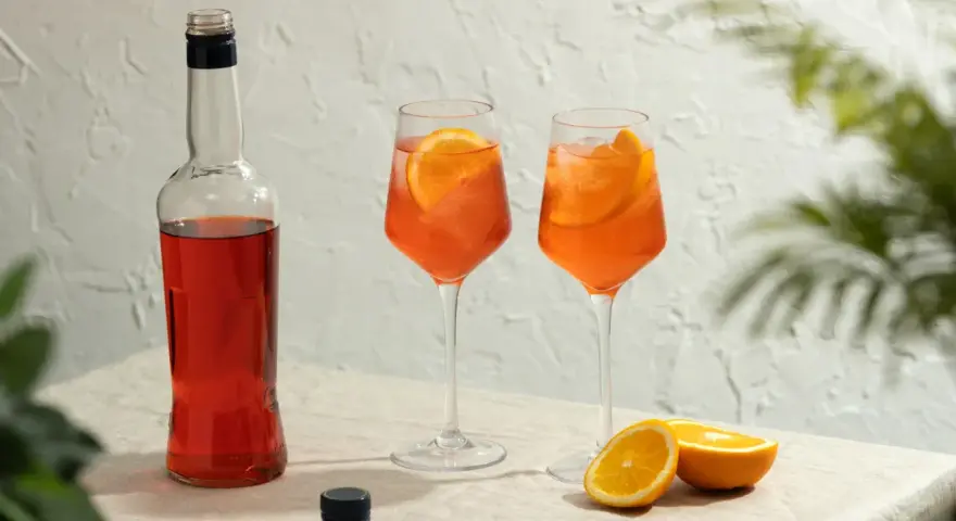 Dwa orzeźwiające drinki aperol z colą, ozdobione plasterkami pomarańczy, stoją obok butelki aperolu.