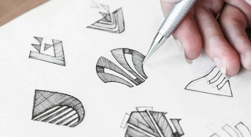 Main d'un designer esquissant des idées pour comment dessiner un logo. Plusieurs croquis abstraits sont visibles sur le papier.