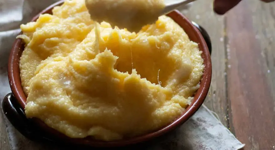 Un cucchiaio solleva una porzione filante di polenta concia valdostana, calda e cremosa, in una ciotola di terracotta.