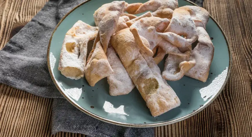 Chrupiące faworki, przygotowane według przepisu babci, posypane cukrem pudrem, leżą na talerzu.