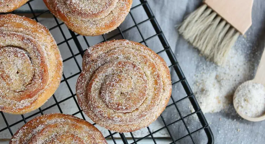Nybakade karlsbaderbullar med socker och kanel på ett svart galler. En borste och en sked med socker ligger bredvid.