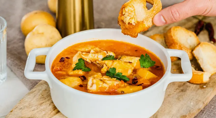 Una mano intinge un crostino nella zuppa di pesce surgelato, ricca di sapori e profumi.