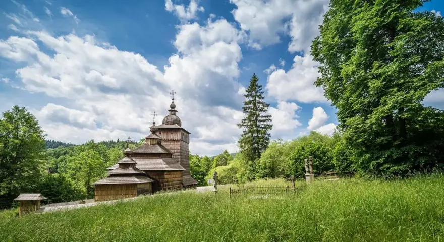 Drewniana cerkiew na tle błękitnego nieba z chmurami, otoczona zielenią. Widok z łąki w Beskidzie Dolnym.