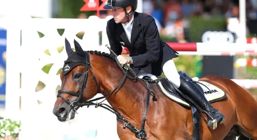 Cavalier en tenue d'équitation, sur un cheval bai, en plein saut. Le cheval, nommé Ratina Z, est concentré.