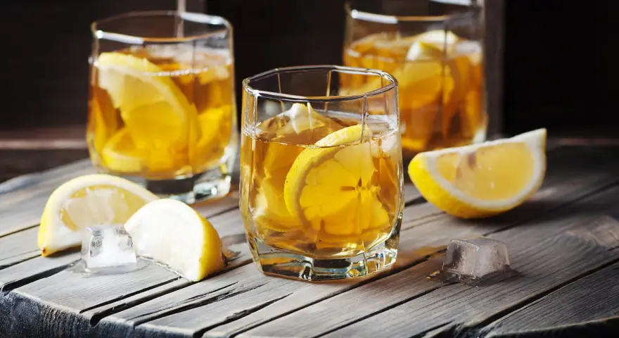 Metaxa orange z czym pić? Idealnie z lodem i plasterkiem cytryny. Trzy szklanki złocistego trunku na drewnianym blacie.