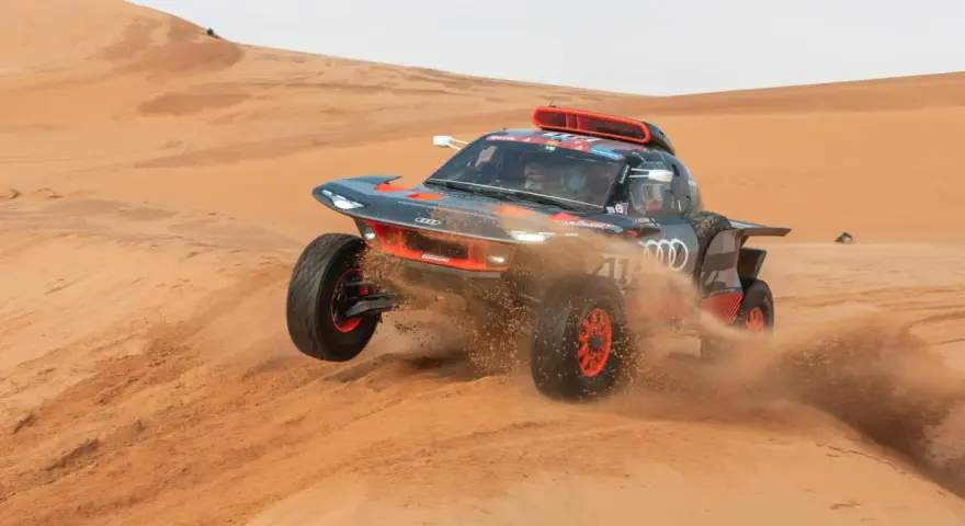 Un'auto da rally Audi sfreccia tra le dune, sollevando sabbia. Un'emozionante tappa del percorso Parigi-Dakar.