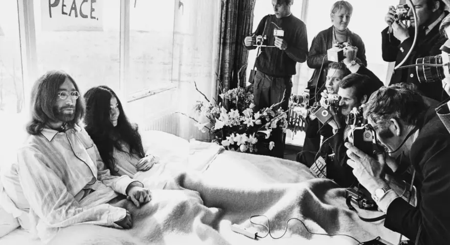 John Lennon i Yoko Ono w łóżku, otoczeni przez reporterów z aparatami. Na ścianie wisi plakat "BED PEACE".