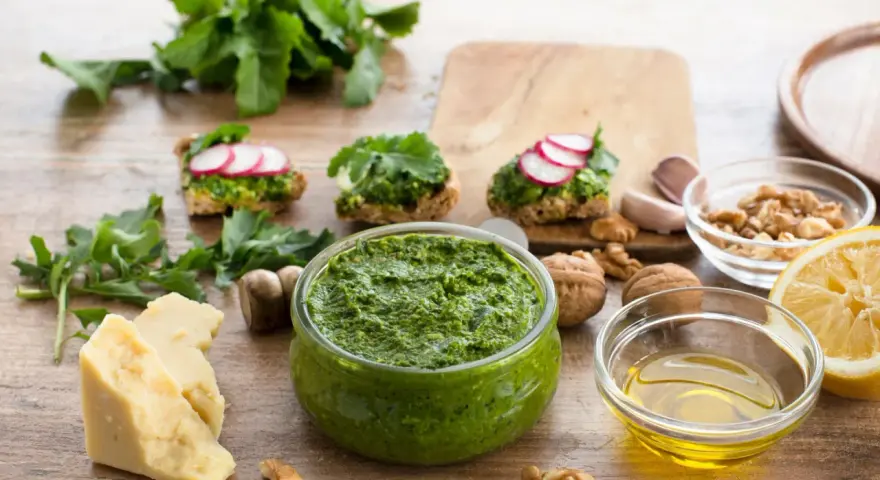 Zielone pesto w misce, obok składniki: ser, orzechy, cytryna, czosnek, oliwa.