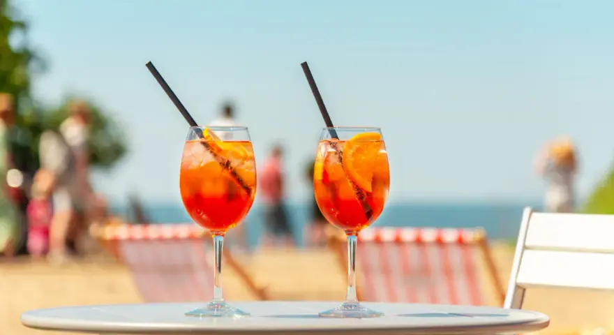 Dwa orzeźwiające drinki aperol z sokiem pomarańczowym, z lodem i plasterkiem pomarańczy, na tle plaży i błękitnego nieba. Idealne na wakacyjny relaks.