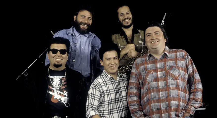 Los Lobos, native sons, pose together. One man wears sunglasses and a fringed jacket, another a denim jacket, and two sport plaid shirts. A saxophone is visible.