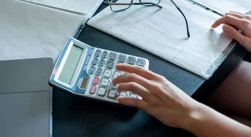 Ręka wpisuje dane na kalkulatorze, przygotowując się do zajęcia wierzytelności. Obok leżą dokumenty i okulary.