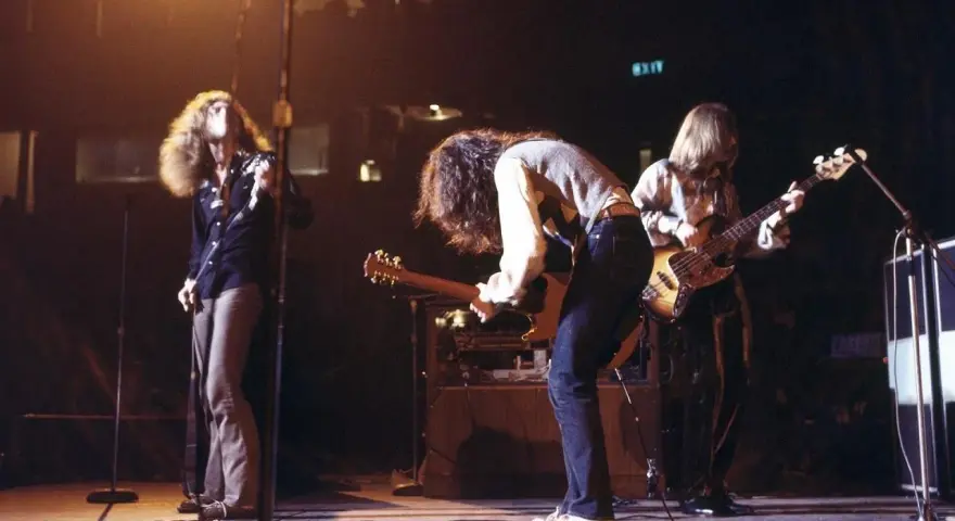 Energetyczny **Led Zeppelin koncert** na scenie. Wokalista z burzą włosów, gitarzysta pochylony nad instrumentem, basista grający w tle.