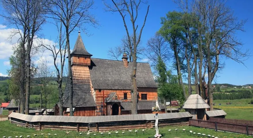 Drewniany kościół w Dębnie, otoczony kamiennym murem i drzewami, z krzyżem misyjnym na trawiastym dziedzińcu.