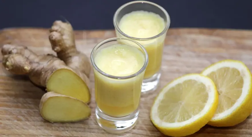 Zwei Gläser mit leuchtend gelbem Ingwer Shot, daneben frischer Ingwer und Zitronenscheiben auf Holzbrett.
