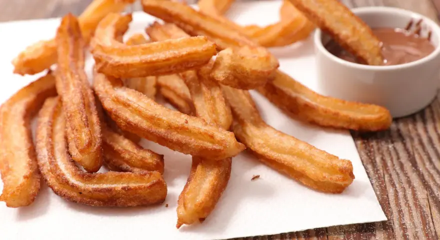 Goldbraune Churros, frisch frittiert, mit Zucker bestreut. Ein Churro wird in eine Schüssel mit Schokoladensauce getunkt. Ein echtes Churros Rezept Original.