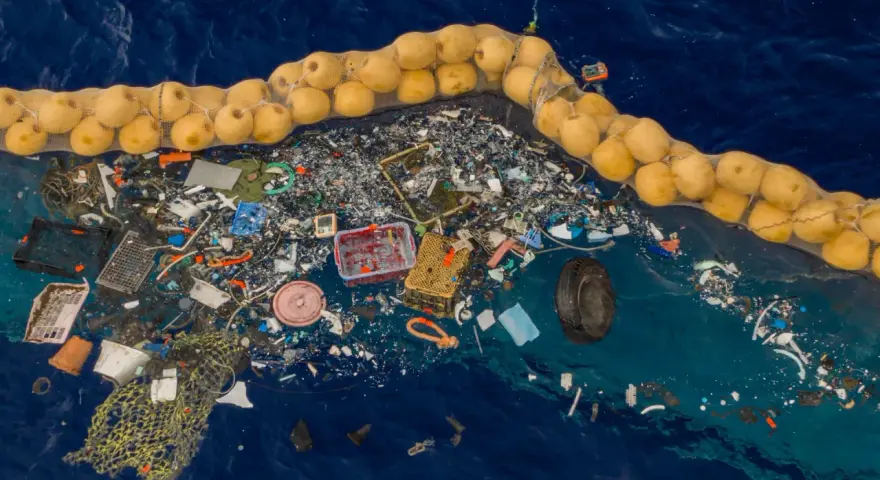 Ogromna wyspa śmieci na oceanie, zebrana w siatkę z bojami. Plastikowe odpady, sieci rybackie i opona tworzą przygnębiający widok.