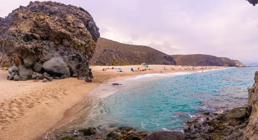 Plaża w Cabo de Gata z turkusową wodą, piaszczystym brzegiem i skalistymi formacjami. Ludzie odpoczywają pod parasolami.