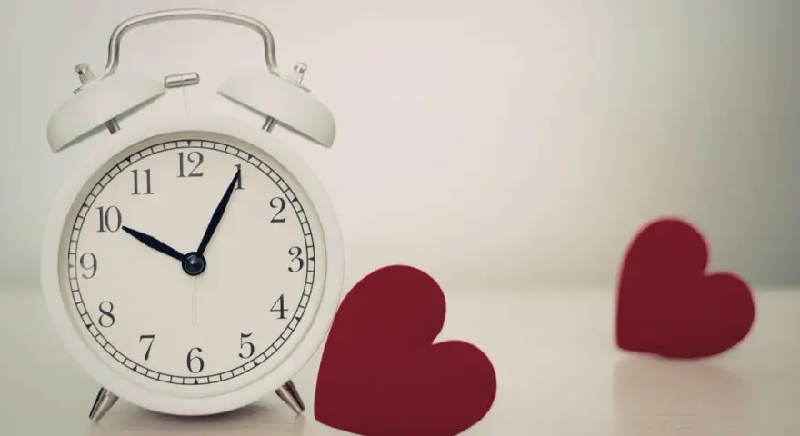 Biały budzik pokazuje 10:08. Obok niego leżą dwa czerwone serca. 0202 znaczenie: czas na miłość.