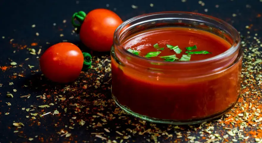 Un vasetto di ketchup fatto in casa, guarnito con basilico fresco, accanto a pomodori maturi e spezie su sfondo scuro.