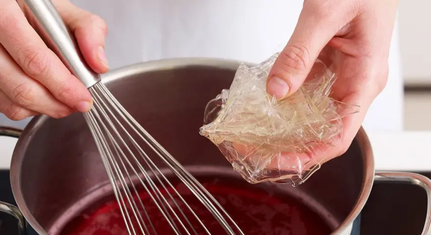Hände bereiten Gelatine-Blätter für die Zubereitung vor, indem sie sie in eine rote Flüssigkeit in einem Topf geben, während ein Schneebesen bereitsteht.