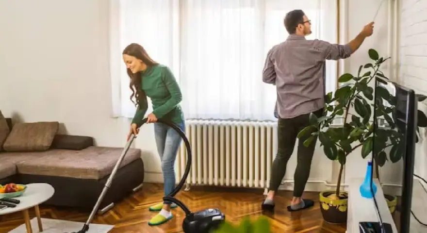 Pareja colaborando para purificar casa. Ella aspira el suelo, él quita el polvo de las paredes.