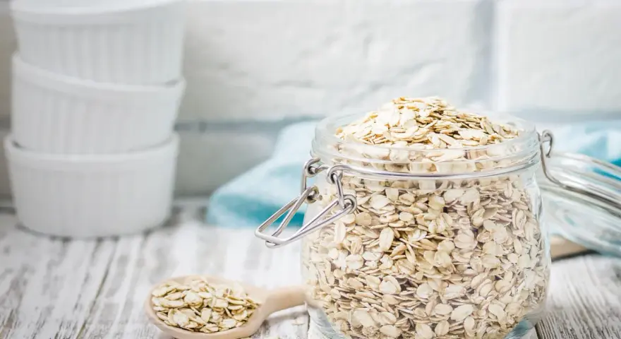 Ein Glas voller haferflocken gemahlen, bereit für ein gesundes Frühstück. Daneben ein Holzlöffel mit weiteren Flocken.