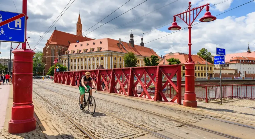Mężczyzna na rowerze jedzie po brukowanej drodze rowerowej z torami tramwajowymi, w tle zabytkowe budynki i czerwony most.