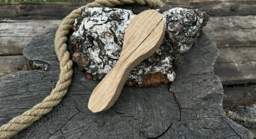 Cucchiaio grezzo in legno d'acero, appoggiato su un tronco scuro con una corda rustica.