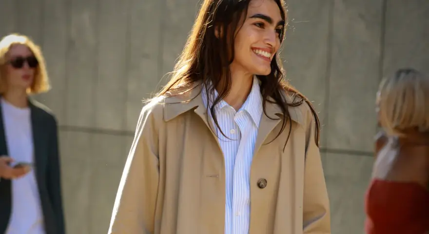 Donna sorridente con capelli lunghi e castani indossa un trench beige aperto su una camicia a righe azzurre. Perfetto per ogni occasione, anche se posso mettere il trench di sera.