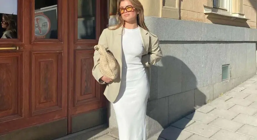 Mujer con blazer y vestido largo blanco, bolso tejido y gafas de sol, posando frente a una puerta de madera.
