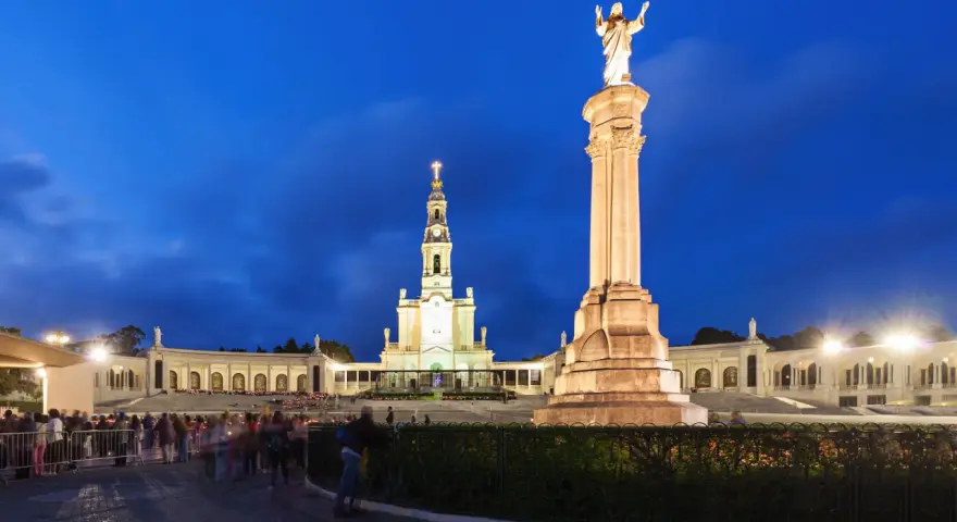 Pilgrims gather at the Sanctuary of Fátima, Portugal, at dusk, with the illuminated basilica and statue of Christ the King standing majestically.