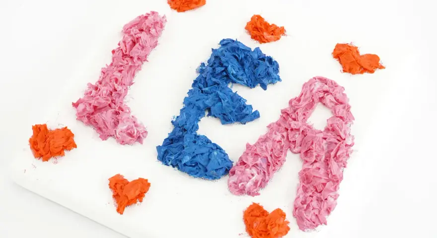 Comment faire du relief sur un tableau : des lettres "LÉA" en papier froissé rose et bleu, entourées de cœurs orange sur une toile blanche.