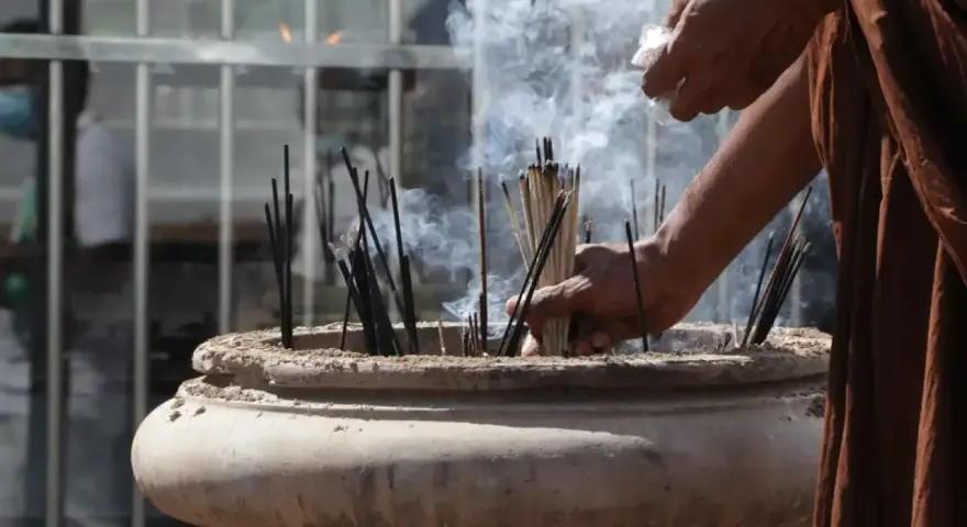 Manos monje en túnica marrón colocando varillas de incienso en un cuenco de piedra, creando humo para limpiar el ambiente.