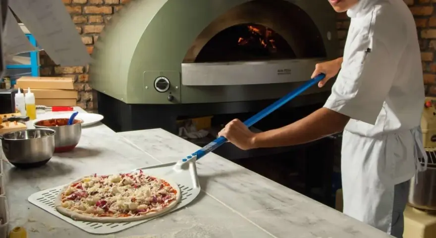 Pizza cruda en pala, lista para horno de leña con chef al fondo.