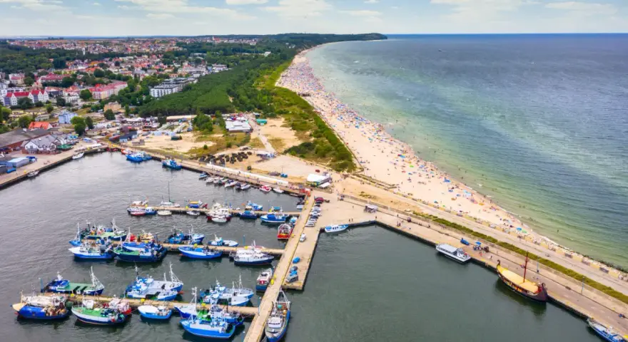 Port rybacki we Władysławowie, województwo pomorskie, tętni życiem. W tle rozciąga się plaża pełna turystów i morze.