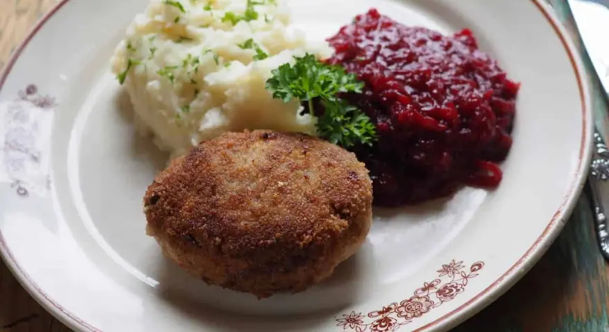 Kotlet mielony z ziemniakami i żurawiną, udekorowany natką pietruszki.