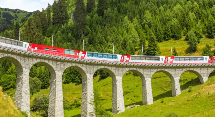 A red and white train travels across a stone viaduct, winding through a lush green valley with a dense forest on the mountainside. This scenic route is part of the Amsterdam to Basel train journey.
